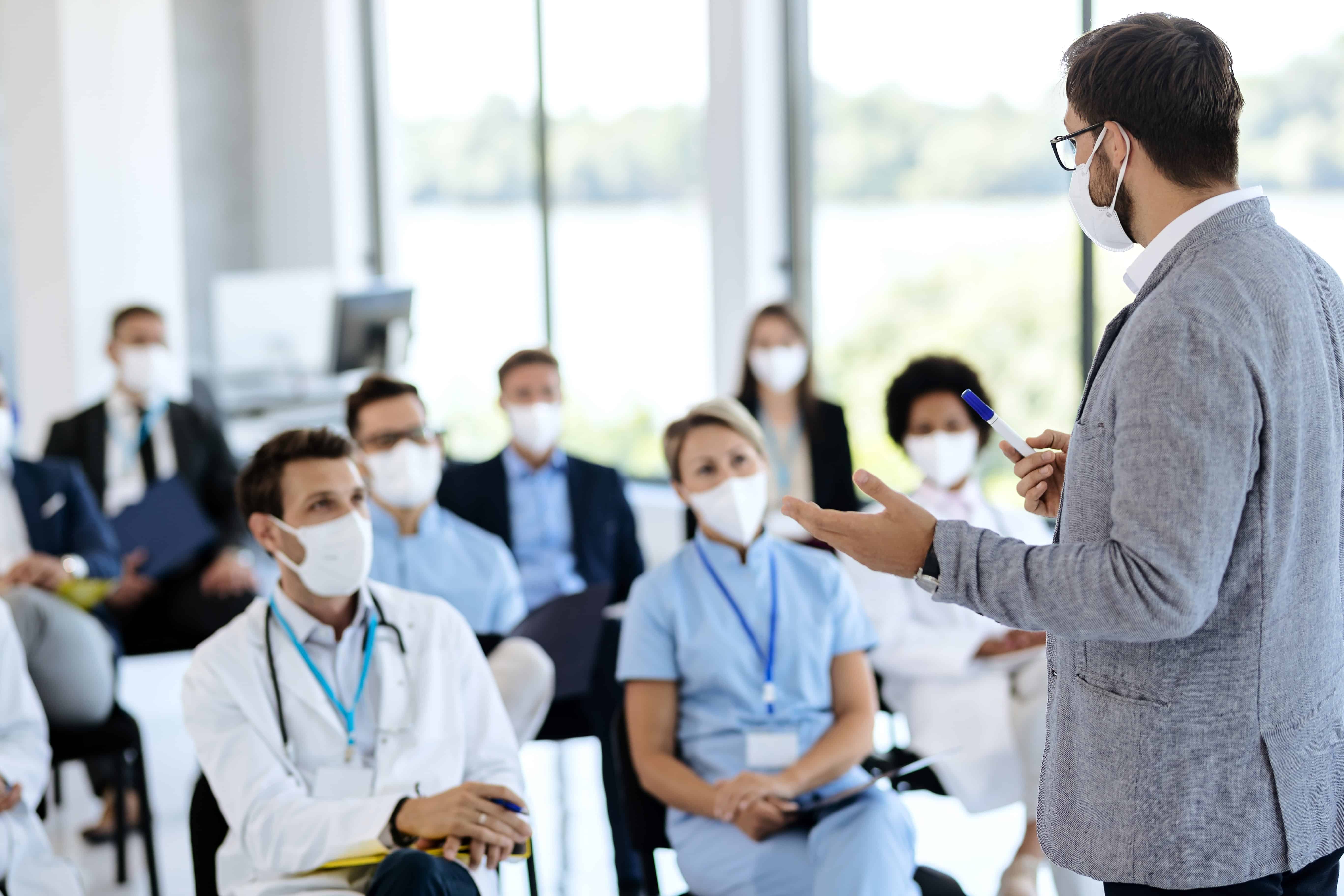 Medical personnel wearing masks and listening to a speaker at an event