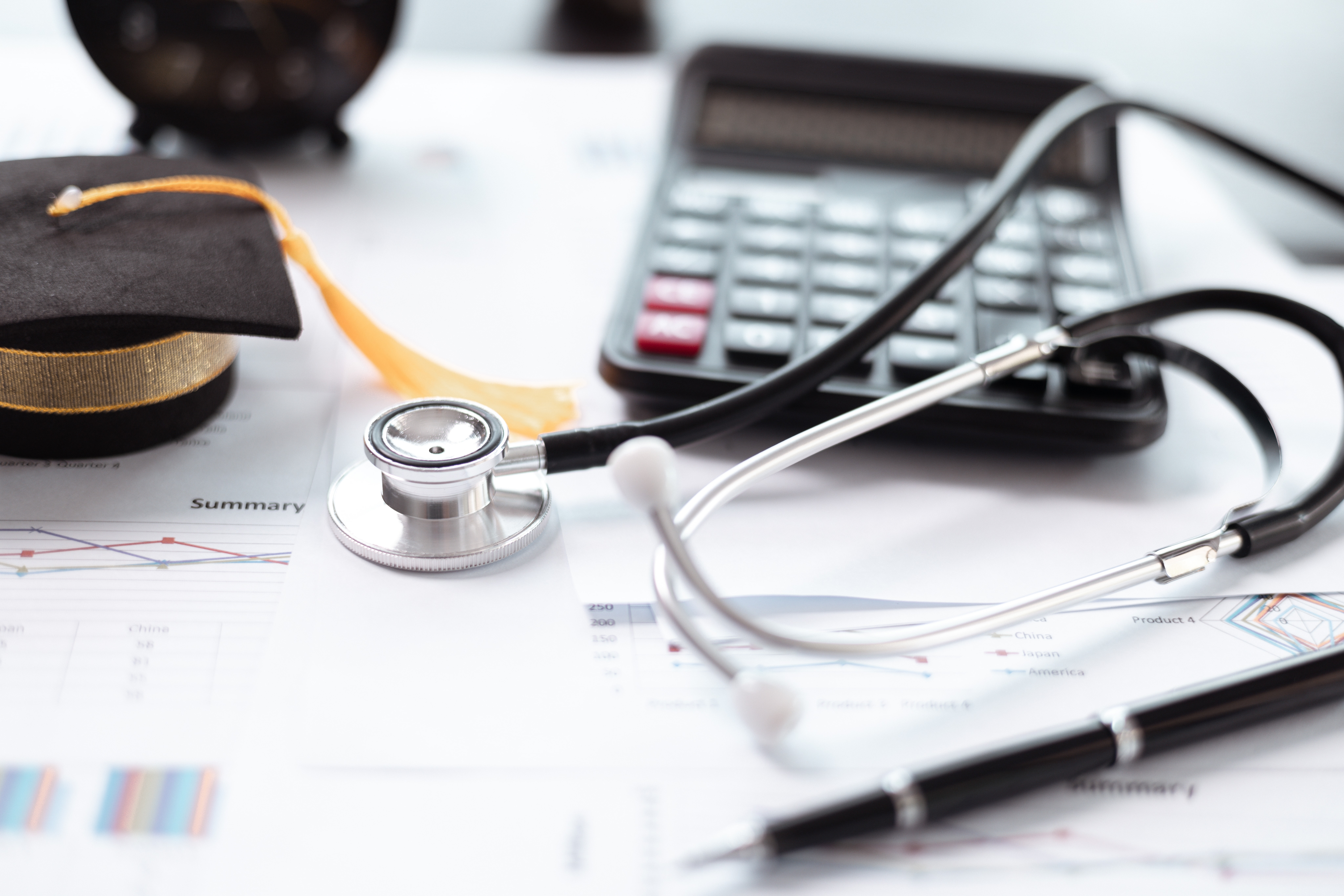 Close up of a calculator, mini graduation cap, stethoscope, and paperwork
